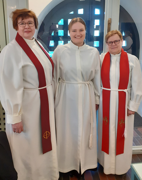 Kolme hymyilevää ihmistä valkoisissa papin alboissa Espoon tuomiokirkossa.